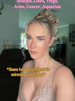 A woman with blonde hair styled up looks directly at the camera with a neutral expression. Text overlays top and bottom, and a gold necklace is visible. The background suggests a living room.