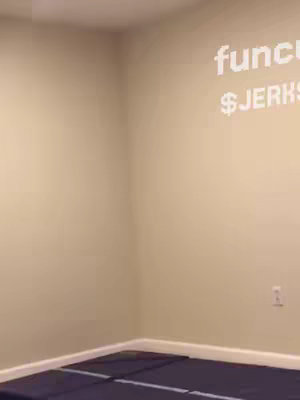 A slightly angled shot of a room corner. The walls are light tan, with white trim at the floor level and at the meeting of the walls and ceiling. A dark blue rectangular object, possibly a mat or a piece of furniture, is on the floor. White text is displayed on the wall.