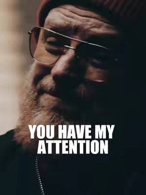 A close-up shot of a man's face. He has a thick, white beard and is wearing glasses and a beanie. The text 'YOU HAVE MY ATTENTION' is superimposed over his face in bold, white font.
