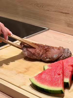 A close-up shot of a wooden cutting board with a grilled steak being handled by a pair of wooden tongs. Sliced watermelon is arranged on the lower right.