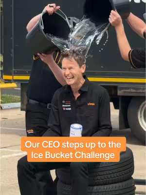 A man smiles widely as water from two black buckets is poured over his head. He's wearing a black short-sleeved shirt and sitting on top of a stack of tires. Another man is pouring one of the buckets and a third man is holding the second bucket. The background includes a black vehicle.