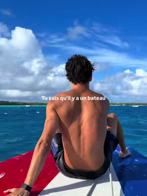 A person sits on a boat, facing away from the camera towards the open ocean. The person has short, dark hair and is shirtless. The sky is blue and filled with puffy clouds. The water is a deep turquoise color.