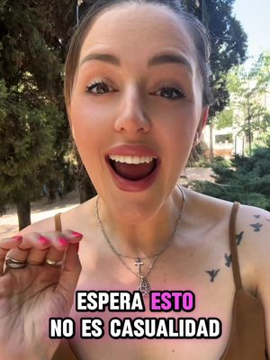 A woman with fair skin and long, light-brown hair and open mouth looks directly into the camera. She is wearing makeup, including eyeshadow and lipstick, and a delicate silver necklace with a cross pendant. A tattoo is visible on her shoulder. Below her, overlaid text reads "ESPERA ESTO NO ES CASUALIDAD". The background suggests an outdoor setting with trees and greenery.