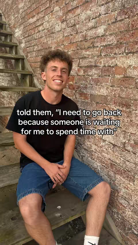 A young man with curly brown hair is smiling and sitting on wooden steps. He's wearing a black t-shirt and denim shorts. The background is a weathered brick wall. Above the man, white text is overlaid on the image.
