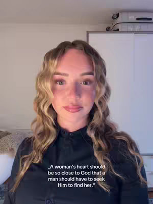 A woman with long, wavy blonde hair is positioned in the center of the frame. She is wearing a black button-up shirt. The background is softly lit, with a glimpse of a curtain or similar fabric on the left and a portion of a white cabinet on the right, with a light fixture peeking from behind her head. The lighting suggests a warm interior ambiance.