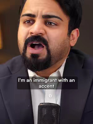 A medium shot of a man with a dark beard and a white shirt, framed from the chest up, speaking into a microphone. A text overlay at the bottom reads, "I'm an immigrant with an accent."
