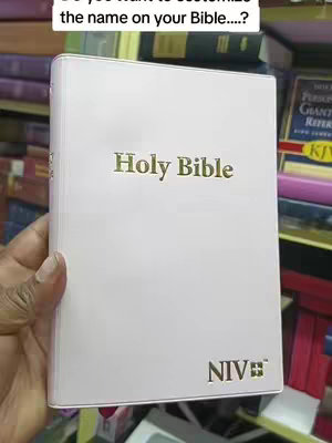 A hand holds a white book that says "Holy Bible" in gold lettering. The bottom of the book has "NIV" in gold. The background is blurred and shows other books.