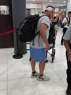 A man with a large backpack and headphones stands in a public space, possibly an airport or convention center, near a wheelchair and another person. The man is wearing shorts and a t-shirt. Another person stands behind him in a wheelchair. The background includes a red rope barrier and signage.