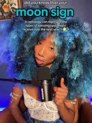 A medium shot of a person with bright blue, voluminous curly hair. In the background, a framed painting with flower imagery is visible. The person is gazing towards the camera, with a slight smile and a nose ring, holding a black microphone with a red and black logo on it. Text overlays in clear, bold fonts are positioned above the person's head. The person is wearing a patterned white top and bracelets.