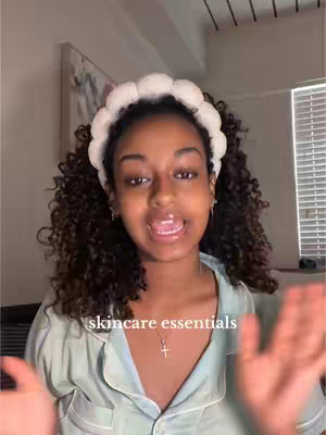 A woman with curly hair and a white headband is centered in the frame, smiling and gesturing with her hands. The background is a muted interior with window blinds and a painting, and text overlayed reads "skincare essentials".