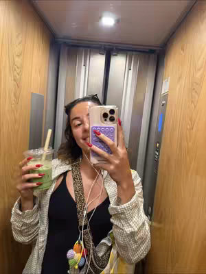A woman is taking a selfie in an elevator. She is holding a green drink in one hand and her phone in the other. She is wearing a black top, a patterned blazer, and has red nail polish. The elevator has wooden paneling.
