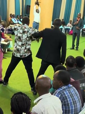 The image shows an active scene inside what appears to be a church or a similar gathering space. The foreground focuses on two men, both seen from the back, walking towards the right. One is dressed in a patterned shirt and black pants, while the other is in a black suit. A third person is standing holding an item over his head behind those men. In the background, a man in a suit is facing the camera with a microphone, and behind him is a seated audience. The scene takes place on a green floor with gold colored walls and blue and white curtains. The overall impression is of a live event, potentially a religious service or a public address.