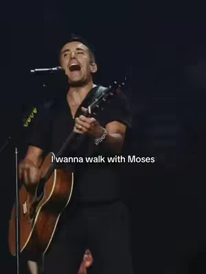 A man is on stage, illuminated by stage lighting, playing an acoustic guitar and singing. The background is dark, and a caption at the bottom says, "I wanna walk with Moses."
