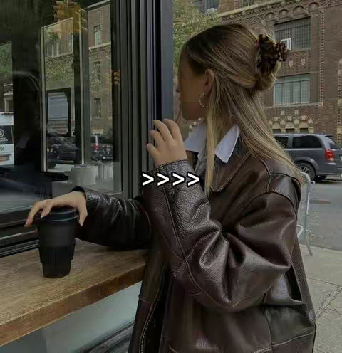 The image shows a woman leaning against a window, outdoors, holding a coffee cup. She is wearing a brown leather jacket and has her hair up. The text overlay consists of four right-pointing chevrons: >>>>>