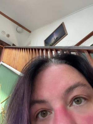 A close-up shot of a person's face with the camera angled upwards. The person's eyes, of a light-brown color, are the focal point. Their skin is fair, with visible redness on the cheeks. Their hair is black with purple highlights. In the background, part of a wooden banister is visible, along with a section of white wall and a framed picture. The overall composition emphasizes the person's proximity to the camera.