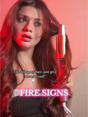 A woman with long red hair is holding a hair curler. The cover image is a medium shot of a woman with red-toned hair and fair skin. She is looking towards the upper left with a slight smile. She is wearing a black top. The background is a gradient of pink to white. Text, "FIRE SIGNS" is superimposed on the bottom of the image.