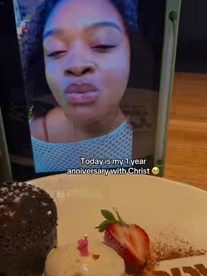 A selfie taken on a tablet shows a dark-haired woman with a plate of dessert in the foreground. The woman has a focused expression, and the tablet displays text indicating a milestone of faith. The dessert plate is set beneath, with a cake, whipped cream, and fruit, suggesting a celebration. The background is blurred, indicating a restaurant or similar setting.