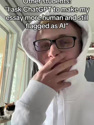 A young man wearing glasses and a white hoodie leans forward, covering his mouth with his hand as if in concern or secrecy. Text overlayed on the image reads a quote about using ChatGPT to make an essay more human, but still getting flagged as AI.