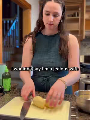 A woman with long brown hair stands in a kitchen, wearing an emerald green sleeveless top. She holds a knife and slices a potato on a white cutting board. The background includes kitchen cabinetry, a window, and a green bottle.