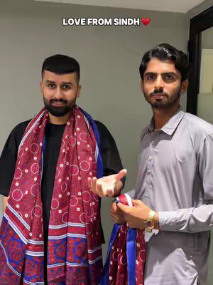 Two men standing side by side, posing in a room. The man on the left wears a black shirt under a red and blue patterned scarf. The other man on the right is wearing a light gray collared shirt and holding a piece of the same scarf.