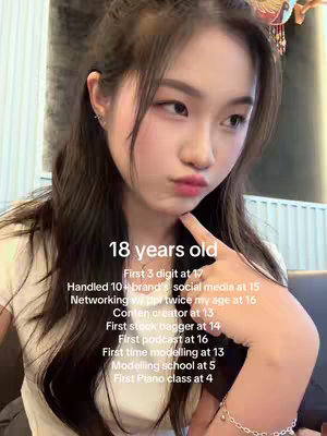 A young woman is the central figure, looking towards the camera with a slight pout. She is holding part of her index finger towards her lips. Overlayed on the image is text detailing her age and personal achievements. The background is blurred, suggesting a potentially indoor setting, possibly a cafe, with a soft, warm light.