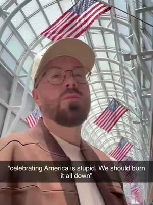 A selfie-style shot featuring a man in a baseball cap and glasses, set against a backdrop of clear, arched ceilings and American flags. The man, with a beard, is in the foreground.