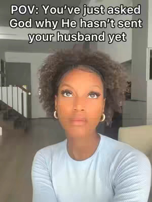 A woman with curly hair and a light blue shirt looks upwards with a questioning expression. The background is a blurred interior of a house with stairs and furniture.
