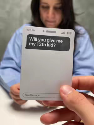 The image shows a person holding a stylized phone-shaped card with a text message bubble. Behind the card, another person is visible, seemingly looking at the card. The card is white with a black text bubble that says 'Will you give me my 13th kid?' showing, along with a new message bar at the bottom. The person is reaching towards the card in the foreground. The background is simple, with a light-colored wall.