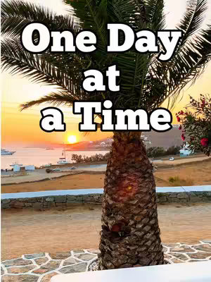The image is a scenic view of a coastal setting, likely near Mykonos, Greece, as indicated by the caption. Centered in the foreground is a textured palm trunk, with its fronds subtly framing the upper portion of the image. Behind it, the horizon features a sunset with a bright sun setting over the sea, the sky glowing with warm yellow and orange tones. The water reflects the sunset's light, and in the middle ground, there are structures and buildings that could be part of a town or village. In the background, the water appears calm. The overall composition is a scenic view, and it conveys a sense of calmness and serenity.