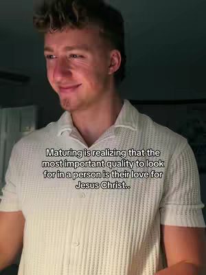 A young man in a white textured button-up shirt is captured in a medium shot, looking slightly off to the side with a gentle smile. Text overlaid on the image offers a religious message. The background is blurred, with indications of an indoor setting, possibly a room.