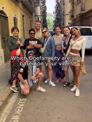 A group of eight people, a mix of men and women, posing for a photo on a city street. They are arranged in a semi-circle. The background shows buildings and narrow streets, indicating an urban setting. Text overlayed on the image reads, 'Which Onefam city are you based on your vibe?' The attire of the people is casual.