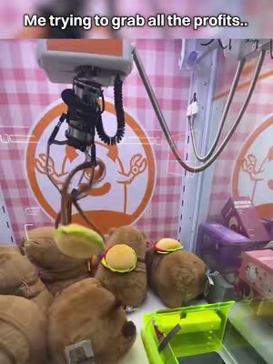 The image shows a claw machine filled with plush toys shaped like brown bears each with a small yellow disc hat. The claw of the machine is reaching downwards towards the toys. The machine is set against a background with a pink and white checked pattern.