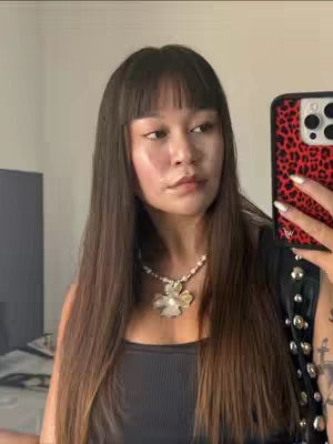 A woman is taking a selfie in a well-lit room. She has long, straight dark brown hair with bangs. She is wearing a black tank top. She is holding a phone up to the side of her face with a red and black leopard print case. She has a pearl necklace with a large, ornate pendant. The background is a neutral color and shows a blurred-out television and a wall.