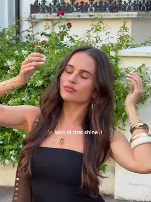 A woman with long brown hair stands outdoors in front of a green bush and a white fence. The image is well-lit with natural sunlight. She is wearing a black top and various gold bracelets and rings.
