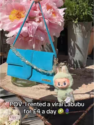 The image shows a turquoise handbag with a sparkling chain, held against a backdrop of pink flowers. A cute Labubu figurine dangles from the bag. The text overlay reads 'POV: I rented a viral Labubu for £4 a day'.