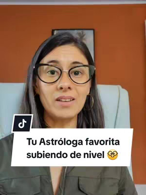 A woman with dark hair and glasses is centered in the frame, looking directly at the camera while talking. Behind her is a muted orange wall with a framed picture, and she is wearing a grey jacket. A white bar at the bottom of the screen contains the text: "Tu Astróloga favorita subiendo de nivel 🤓🤭".
