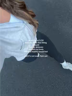 A selfie-style video shows a person from the chest down, angled slightly from above and to the side. The person is wearing a white t-shirt over a blue shirt and black leggings. The background appears to be a dark-colored surface. Text is overlaid on the image, detailing a morning routine. The overall composition is simple, with a focus on the person and the text overlay.