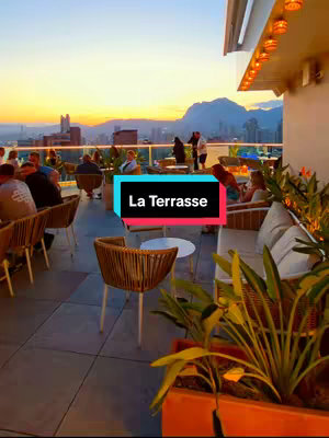 A rooftop bar at sunset with a view of a city and mountains in the background. The image has a vertical orientation and features a foreground of planted greenery and decorative seating, with patrons visible throughout. The sky is a gradient of orange and blue hues, indicating sunset.