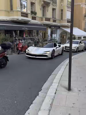 A street scene in Monaco featuring a white sports car, likely a Ferrari, driving on a cobblestone street. The background includes buildings, shops, and parked vehicles.