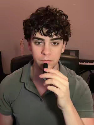 A young man with curly dark hair stands in front of a dark background, holding a small black object near his mouth. He's wearing a green polo shirt and looking towards the camera with a serious expression.