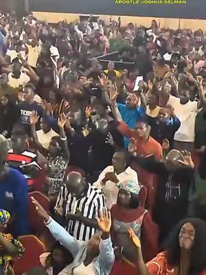 A crowd of people with their hands raised in worship during a religious service. The focus is on the congregation, with the speaker, Apostle Joshua Selman, visible in the foreground. The lighting is bright, presumably from overhead sources, and the atmosphere seems energetic and spiritual.