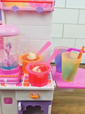 The image shows a pink and white toy kitchen set-up with various colorful desserts and drinks in small clear containers and cups. A blender dominates the left side, and several dessert samples in vibrant shades of red and orange are displayed on shelves. A sample drink is on the right in an ombre mix of green and yellow. The composition is an inviting display of a children's toy set.