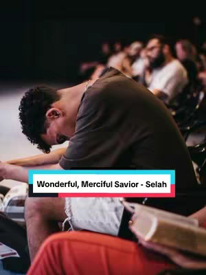 A man with short, dark hair and wearing a dark t-shirt is the central focus, leaning forward with his head bowed, seemingly in a moment of worship or contemplation. He is seated among other individuals, only partially visible, in what seems to be an auditorium or church setting. A rectangular text overlay at the bottom features the title of a song.