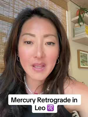 A woman with brown hair and a white necklace is speaking to the camera. The background is a light wooden blinds and a bookshelf or desk. A text overlay at the bottom reads "Mercury Retrograde in Leo" with the Leo astrological symbol.