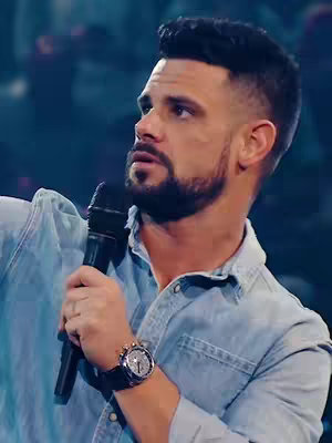 The image is a medium shot capturing a man with a short, dark, neatly styled haircut and a well-groomed beard, set against a blurred, dark background that suggests a stage or auditorium. The man is holding a microphone in his right hand and looking up and slightly to his left with a serious expression. He's wearing a light blue denim shirt with the sleeves rolled up to his elbows, and a watch with a silver face and black strap. The overall lighting appears to be a mix of natural and stage lighting, enhancing shadows and defining his features. The composition and his eye line suggest a speaker or performer in the middle of a presentation or speech.