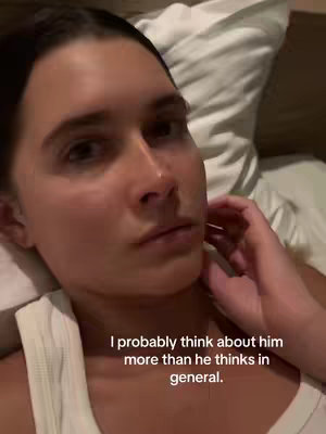 A close-up selfie of a woman lying in bed. She is looking up and slightly to the left, with a thoughtful expression. A white pillow is behind her, and some of her arm is visible.