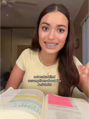 A young woman with long brown hair is seated at a table, looking directly at the camera and smiling. She wears a light yellow t-shirt and braces. An open book with notes and a pink sticky note is in front of her. The background includes a ceiling fan and a glimpse of a room.