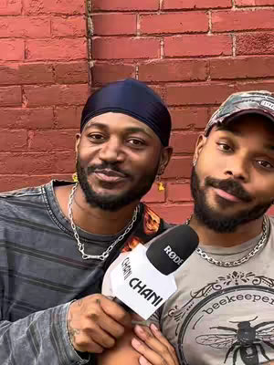 Two men are positioned close together in front of a red brick wall. The man on the left wears a dark durag and a grey t-shirt, with a silver chain necklace. The man on the right is wearing a camouflage cap, silver chain, and a t-shirt that partially obscures the logo in front. Both have short, dark facial hair.