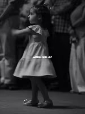 A grayscale image captures a young girl in a flowing dress, dancing amidst blurred figures. The foreground features the girl, mid-movement, with an arm extended and a slight smile. Text overlayed in white reads 'WITH OUR HANDS'. The background is softly focused, with indistinct figures suggesting a crowd or gathering. Lighting seems natural, with diffused shadows, creating a candid feel.
