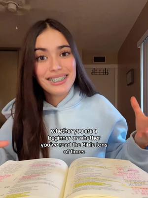 A young woman with long dark hair and braces on her teeth is smiling at the camera, with her arms outstretched slightly. She is wearing a light blue hoodie. In front of her, there's an open Bible with highlighted text and notes visible. Text overlayed in white font states, 'whether you are a beginner or whether you've read the Bible tons of times.' The background features a wall, door and a window.